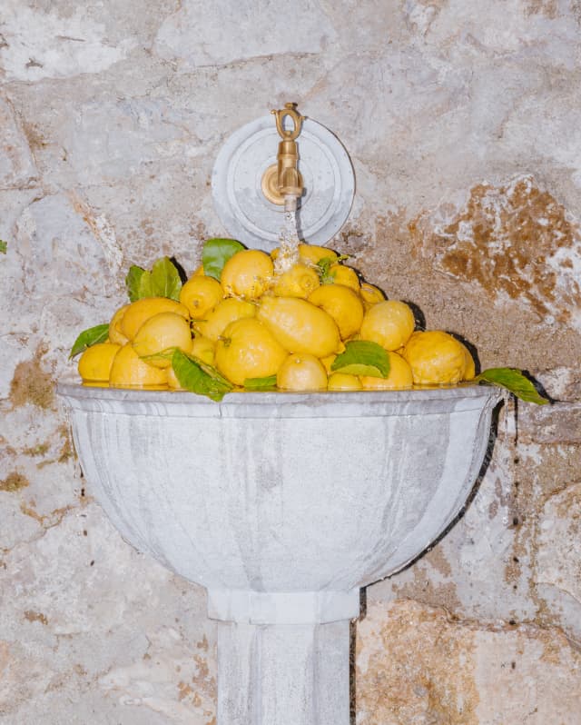 A mountain of lemons fills a marble sink set into a stone exterior wall as a running  tap overflows