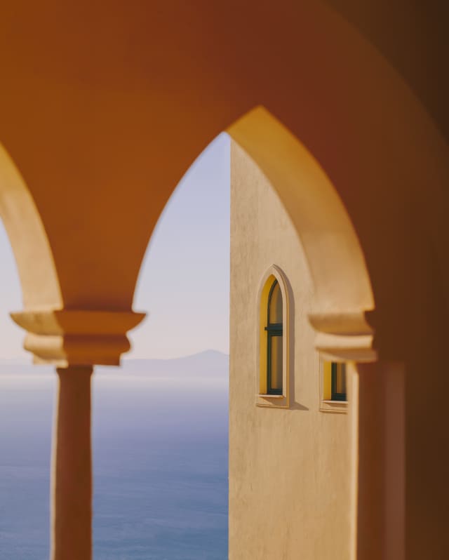 A pointed Moorish arch in Naples yellow is turned honey gold by the low sun as it reveals ocean views of deepest azure