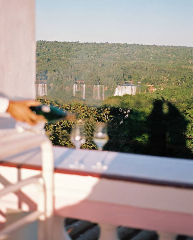 In soft-focus, two glasses on the terrace balustrade receive a top up of sparkling wine to be enjoyed with waterfall views.
