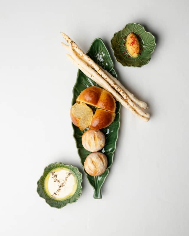 Three majolica leaf dishes with two cornbread buns, a quartered brioche, fresh milk curd and butter, served at Restaurant Y.