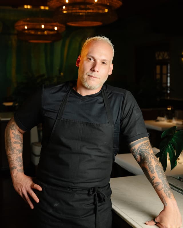 Restaurant Y Chef Luiz Filipe Souza, blond with tattoos, wears a black shirt and apron and leans on a table with one arm.