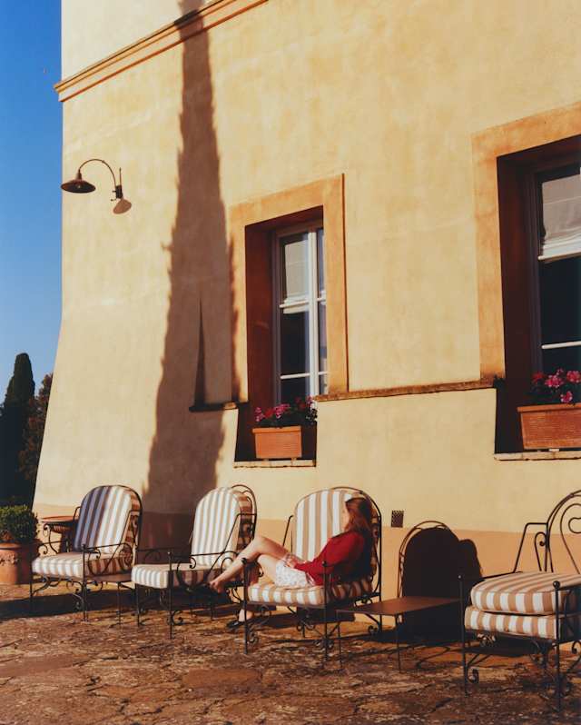 A woman relaxes in one of four patio chairs by a sunny exterior wall brushed by the tall shadow of a cypress tree off camera.