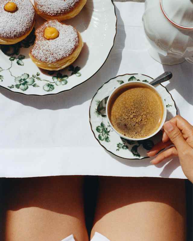 A woman holds the handle of her coffee cup at a table with a plate of three sugar-dusted bombolones, seen from above.
