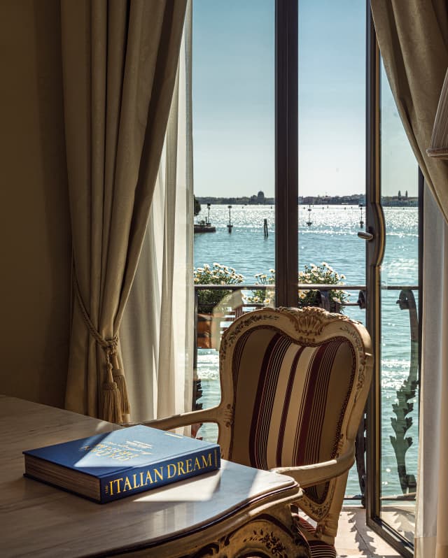 A blue hardback Italian Dream, its title embossed in gold, sits beside an antique chair. Through windows, sun dances on water