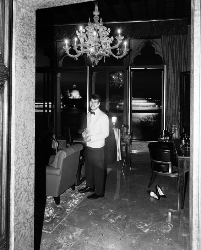 A waiter in a white jacket smiles to the camera as he serves wine in a private lounge below a Murano glass chandelier