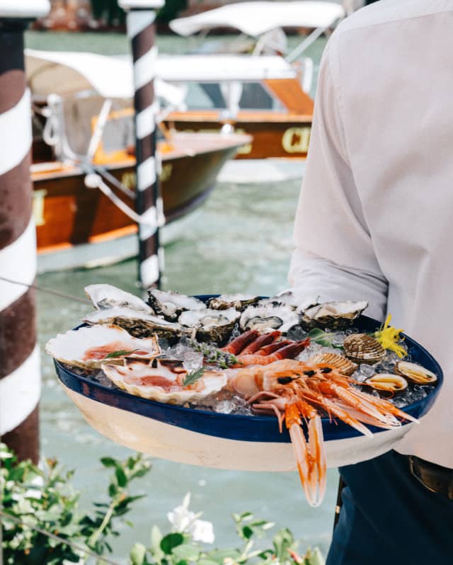 A waiter carries an iced seafood platter. Behind him the Hotel Cipriani’s private launch is moored to one of the paline de casade