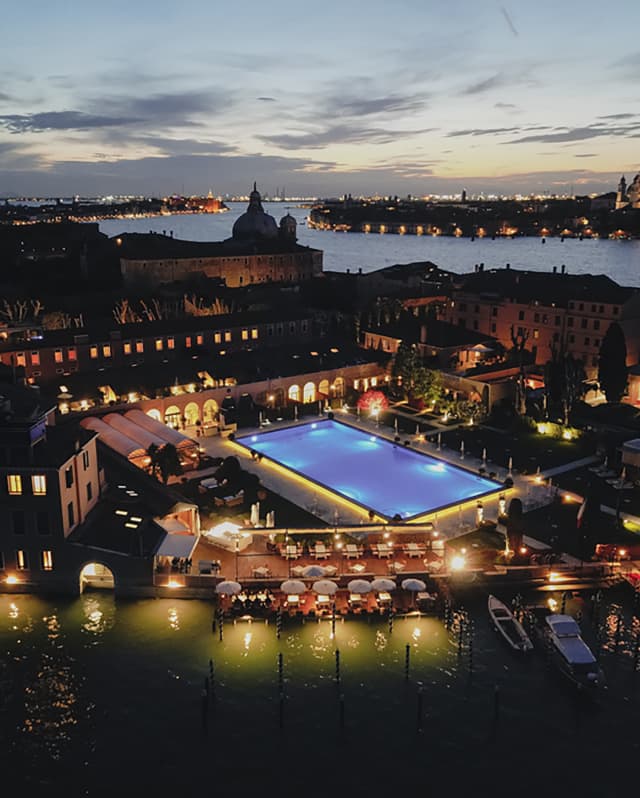 Aerial night view of Hotel Cipriani, sublit pool glowing blue and lights twinkling on Venice's docks, homes and landmarks.