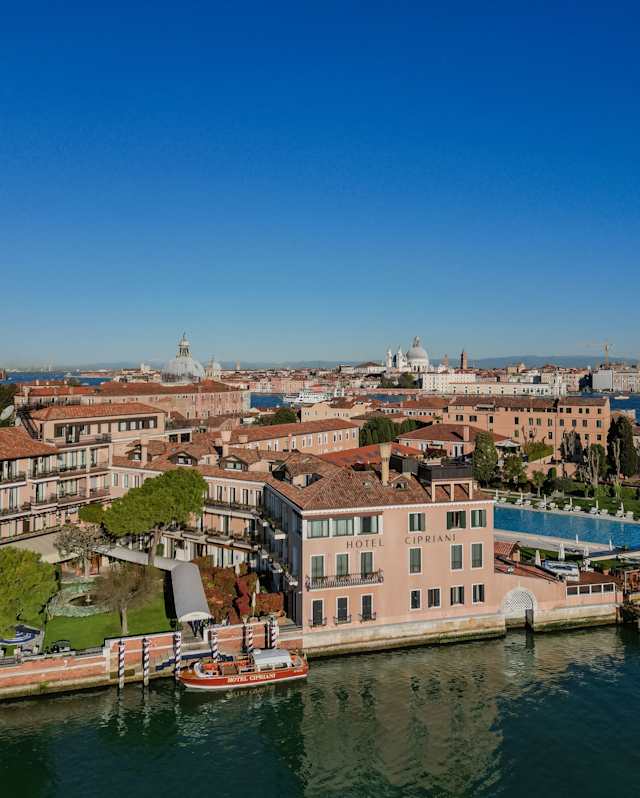 Aerial view of Hotel Cipriani's front, where a boat waits by striped mooring posts, and glimpses of greater Venice beyond.