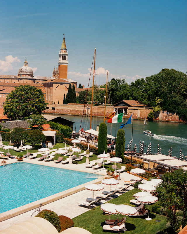 High-angle view over the pool, lined with parasols, and the canal, to the park, dome and clocktower of San Giorgio Maggiore.