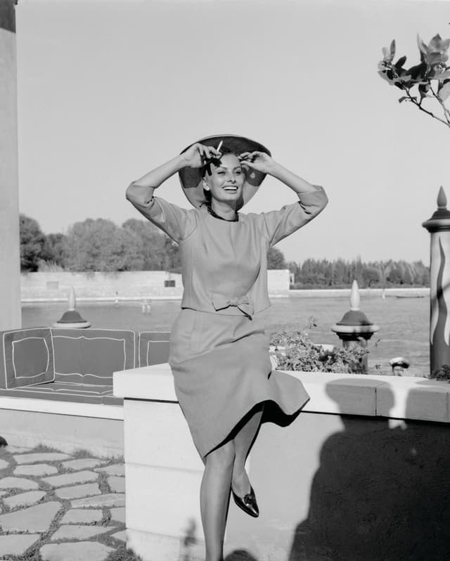 A black and white image of Italian actress, Sophia Loren, smiling as she sits on a low wall in grounds of the Hotel Cipriani