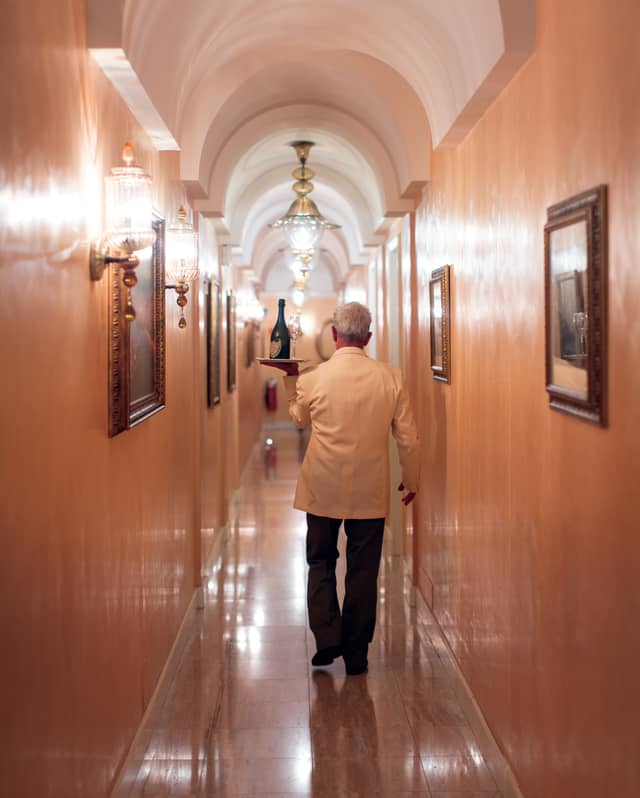 A waiter carries aloft a silver tray of wine and glasses for room service, through a vaulted corridor lit by Murano glass lights