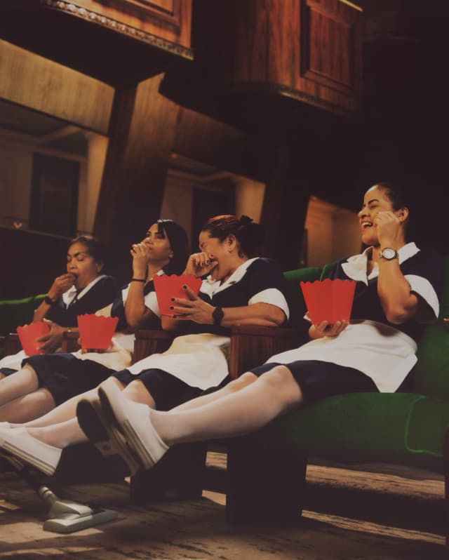 Four uniformed staff laugh and eat popcorn on the green theatre chairs, lifting their legs for a cleaner's vacuum to pass.