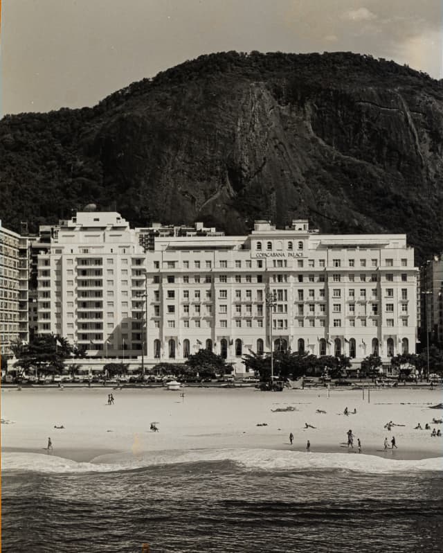 A vintage image of the hotel's facade and beach with the forested mountain behind shows almost no difference to today