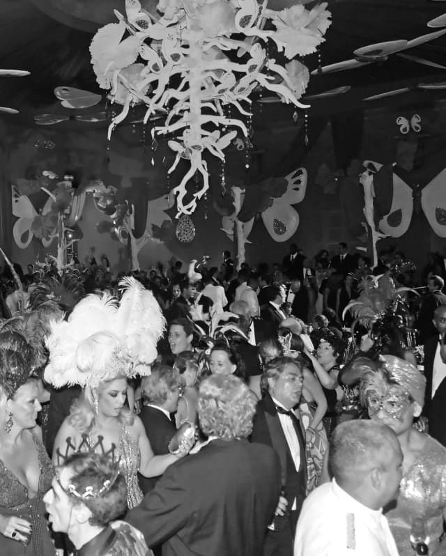 Giant butterflies hang from the ceiling and walls. Party-goers in carnival costumes and black tie fill the ballroom and dance