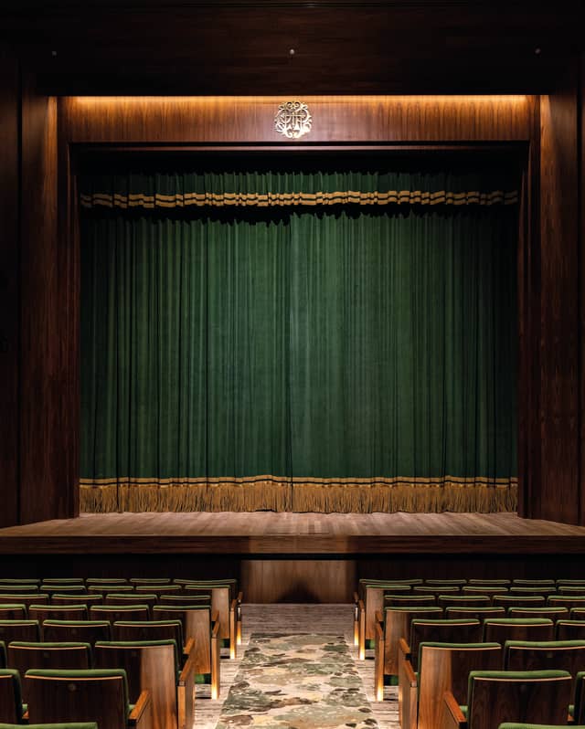 View down the aisle of the dimly-lit theatre, flanked by green stall seats, leading to the stage with a green safety curtain.