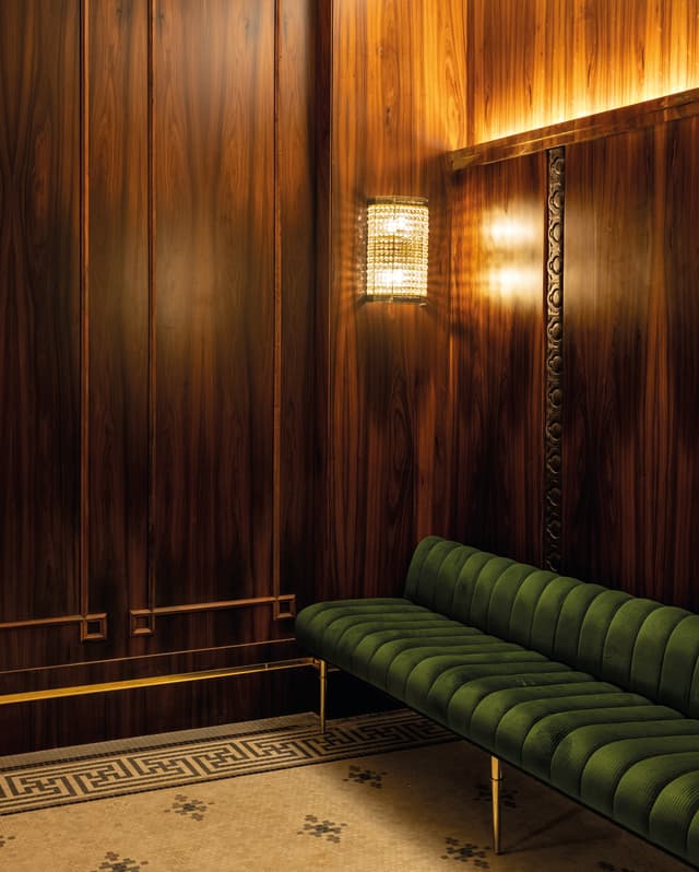 A velvet green bench hugs the length of a wood panelled wall. Art deco patterns are etched in the panels