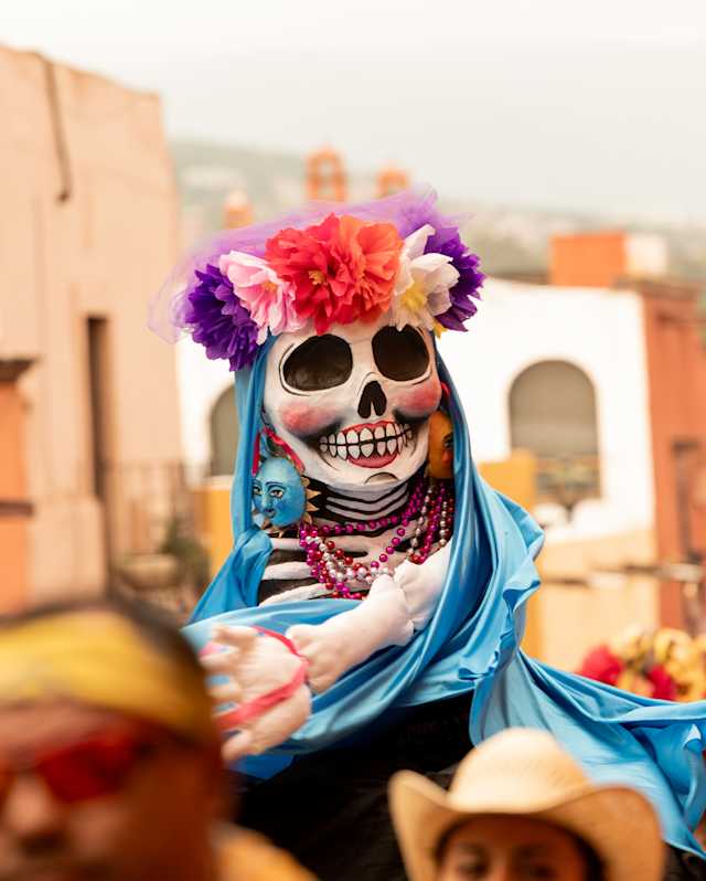 A skull-faced puppet wearing a red, pink and purple floral headband and blue cloak is held above crowds in a street parade.