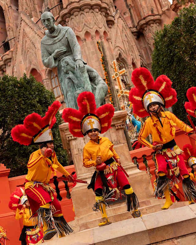 Angled capture of La Alborada dancers as they perform in huge red flowerlike headpieces with yellow shirts and knee tassels.