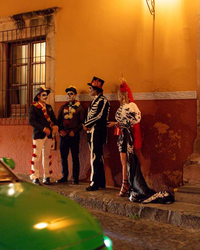 A woman and three men stand beneath a glowing street lamp in Dia de Muertos costumes with skull-painted faces.
