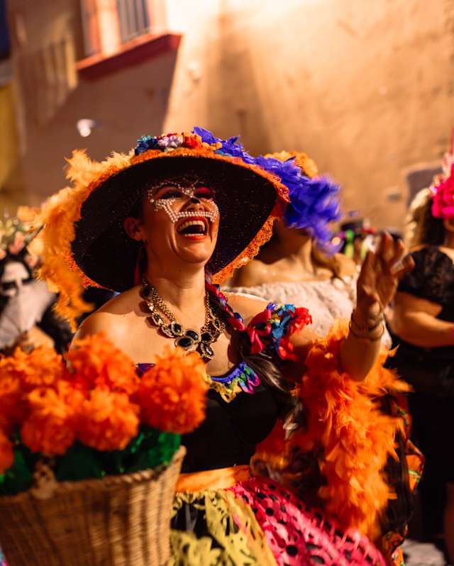 A woman in a flamboyant skirt and hat in pinks and purples, with marigold-orange feathers, enjoys the Dia de Muertos parade.