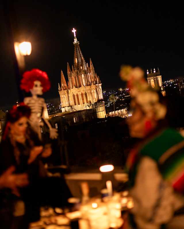San Miguel's parroquia glows against the night sky, seen from a rooftop view over Dia de Muertos decorations in soft-focus.