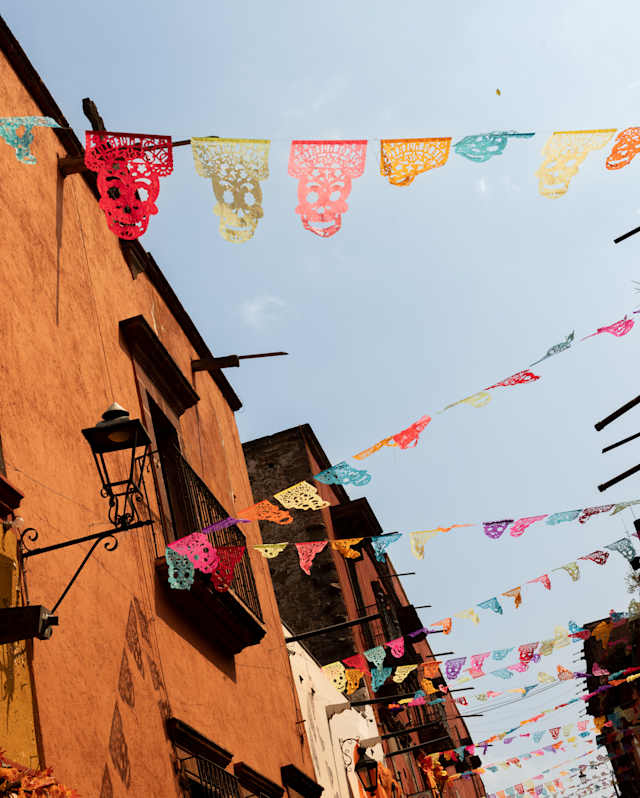 Strings of bright papel picado zigzag between the orange-rendered buildings of a San Miguel street, seen from below.