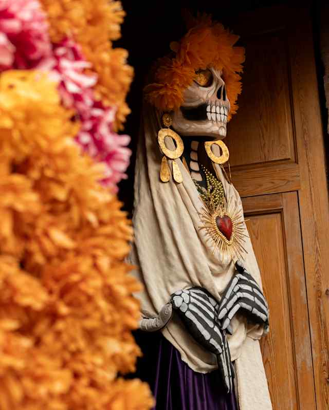A Dia de Muertos skull-head figure stands in a doorway in large earrings and a necklace, part-obscured by a marigold garland.
