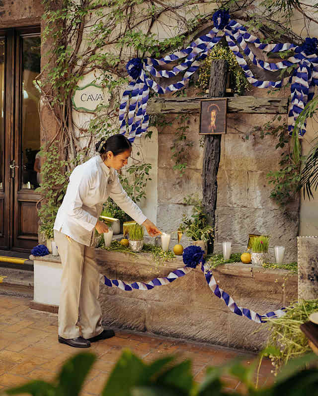 A woman lights a candles at a wall shrine with a wooden cross and a religious picture adorned with a blue and white ribbon.