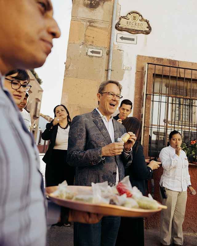 A man in a black suit and glasses unwraps an offering served in the street at a festival, seen over a tray of wrapped sweets.