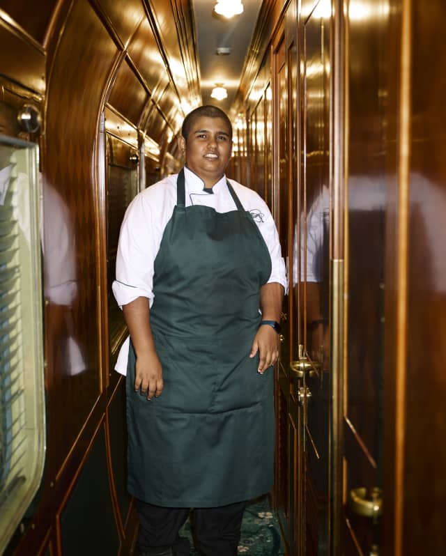 A staff member in a green apron stands beneath lights in a corridor of gleaming polished wood, in a candid shot by Kitajima.​