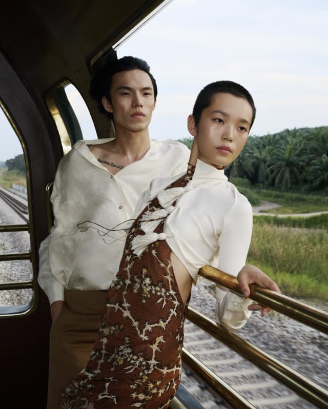 A woman in a brown floral-print dress and white bolero top leans back on the Observation Car rails, alongside a male friend.