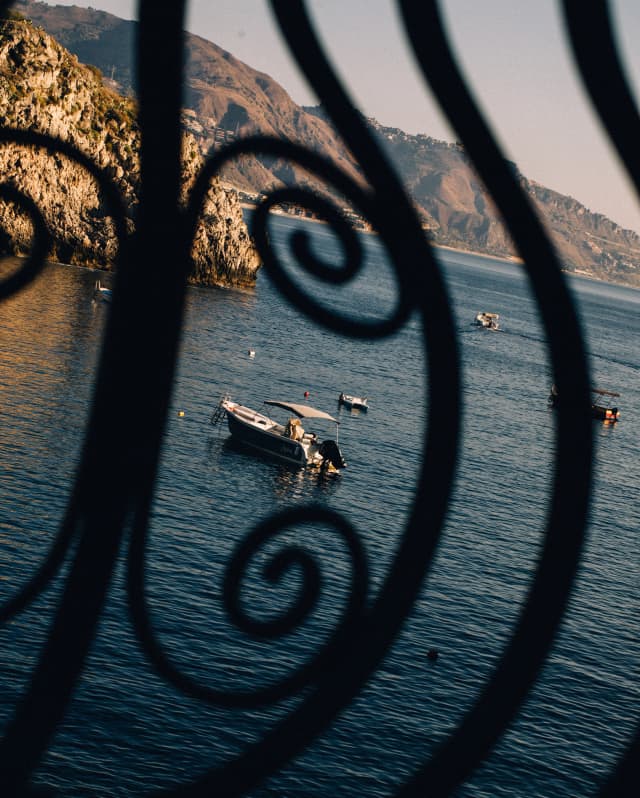 A boat floats on calm sea near a rocky headland, with others further out, glimpsed through curled wrought iron railings.