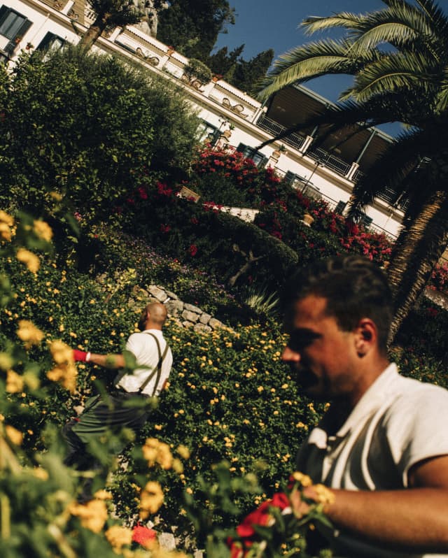 Angled image of the hotel gardens, where two gardeners in white shirts and red gloves attend to the yellow flowering shrubs.