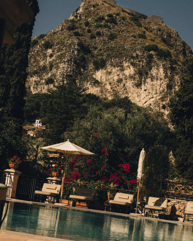 Ground-level view over the pool, where loungers recline beneath two parasols, one folded, with a dramatic mountain backdrop.