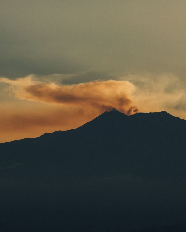 Dramatic image of smoke rising from the silhouetted Mount Etna, melting with the sunset to create plumes of orange and brown.