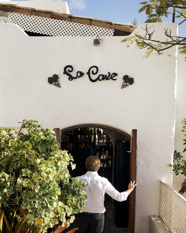 A man in black trousers and a white shirt opens the double doors to the La Cave cellars and tasting rooms, seen from behind.