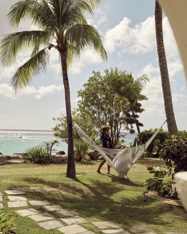 A woman in a hat chats with woman in white who sits on a hammock strung between two palms in a green garden by the ocean.