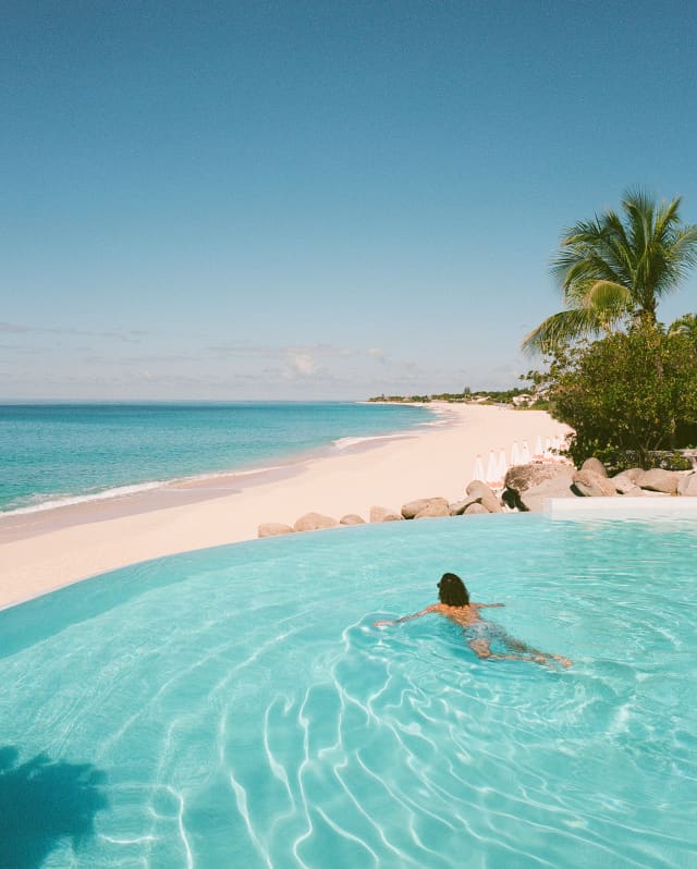 Divided by a strip of white beach, the azure sea complements the bright water of the infinity pool where a man swims alone.