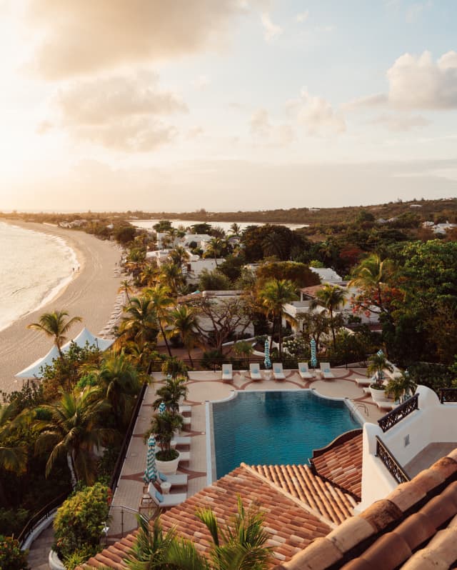 Sunset glows on the sand, roof tiles, palm trees and pool in a birds-eye view of the resort and distant sweep of Baie Longue.