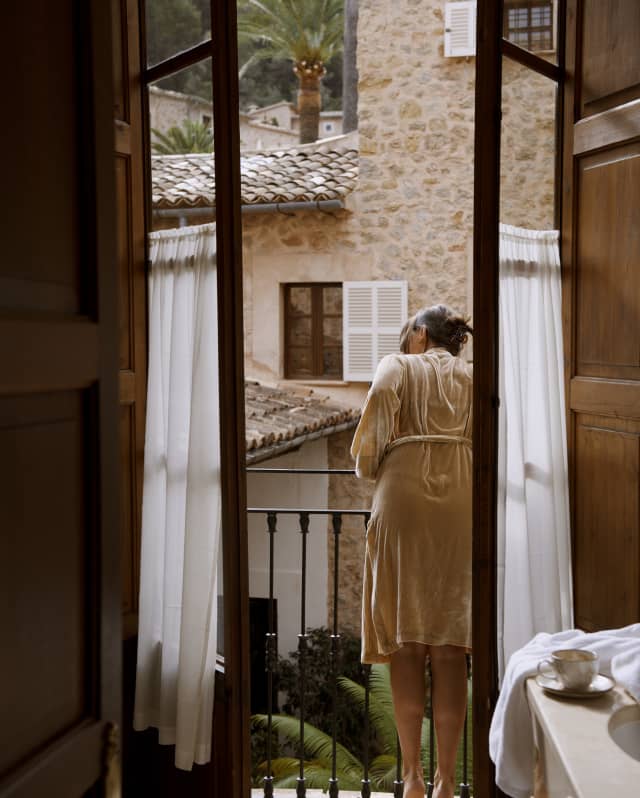 A woman in a gown leans over her balcony to see what's happening in the honey-stoned street below, viewed through open doors.