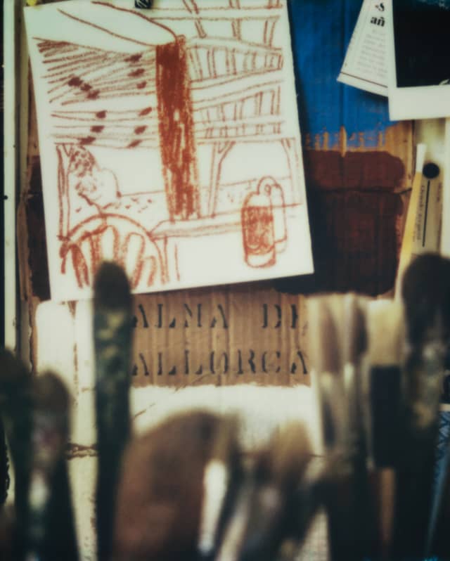 Sketches and images are pinned to an artist's easel. In the foreground, out-of-focus paintbrushes stand like tall grasses