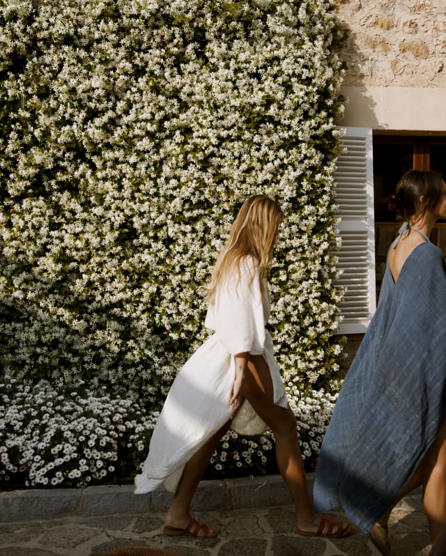 A female guest with long blond hair, wearing a white linen wrap dress strides past the jasmine-clad hotel, seen side on.