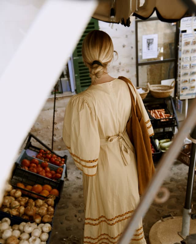 Seen from behind through a canopy, a blond woman in a white summer dress browses a vegetable stall in a corner of Deià.