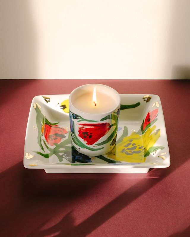 A lit candle with colourful floral designs sits on a matching square ceramic dish, both featuring painted red, yellow, and green patterns, placed on a red surface with a shadow in the background.