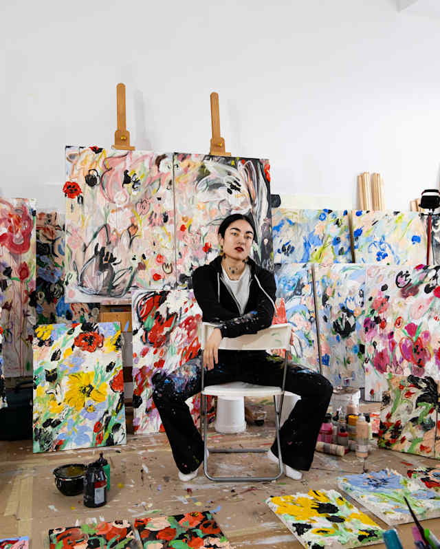 An artist sits on a stool in a studio, surrounded by numerous colourful abstract paintings on canvases and scattered on the floor. Paint supplies are visible in the foreground. The room is bright with white walls.