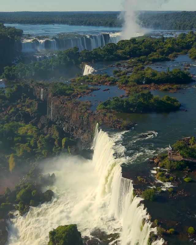 Aerial view of the Iguassu falls with creamy rapids cascading over the forested rocky edge, with more falls in the background.