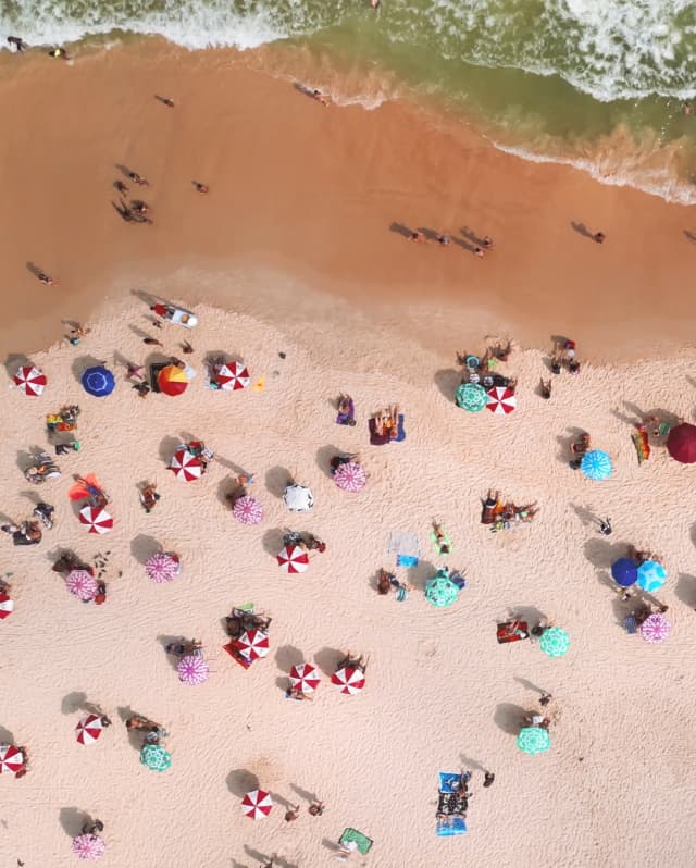 Aerial shot of a pinkish sand beach dotted with sunbathers and candy-like parasols in mint, pink, and red-and-white stripes.