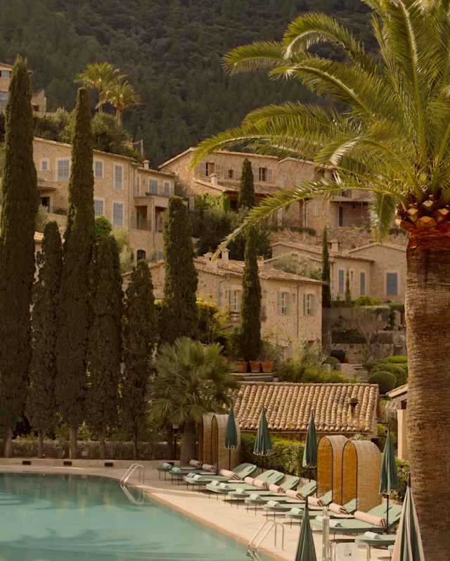 A serene outdoor swimming pool with loungers and umbrellas is surrounded by tall palm trees and cypress trees, set against a hillside with rustic stone buildings and lush greenery.