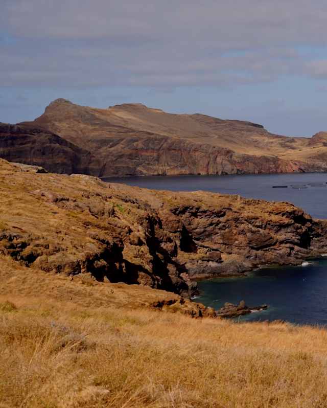 A coastal landscape with rugged, brown hills and cliffs meeting a calm blue sea under a partly cloudy sky. The foreground is covered in dry, golden grass.
