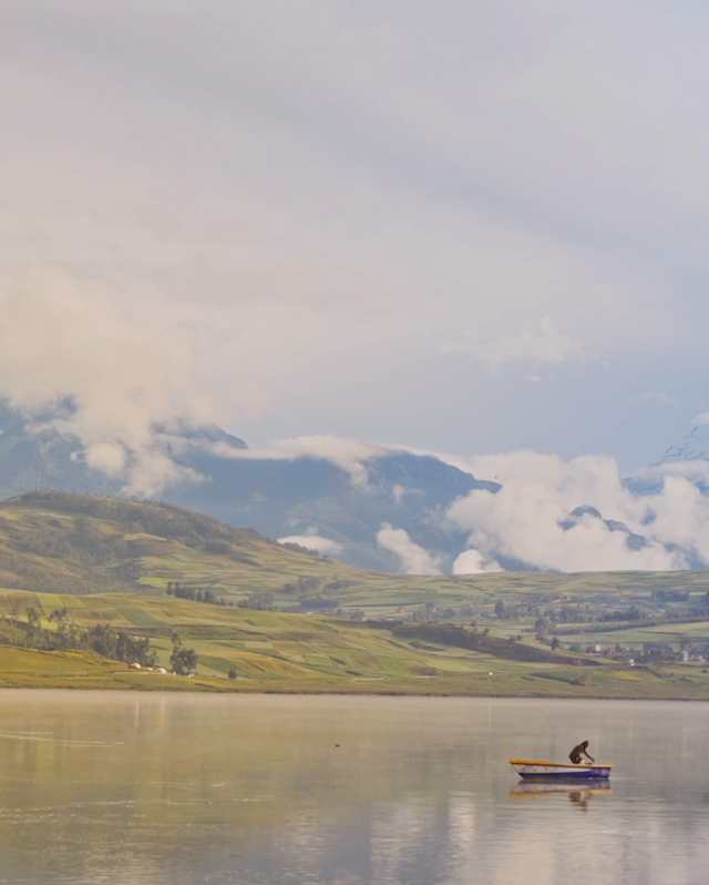 Snowy peaks rise on the horizon in a view over Laguna Huaypo, where a man tends to a boat on the still, green-tinted waters.